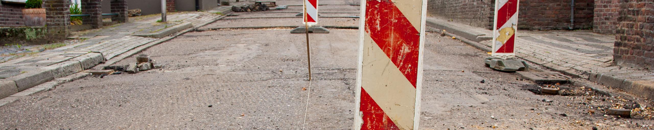 Opengebroken straat met putdeksel en rood witte paaltjes